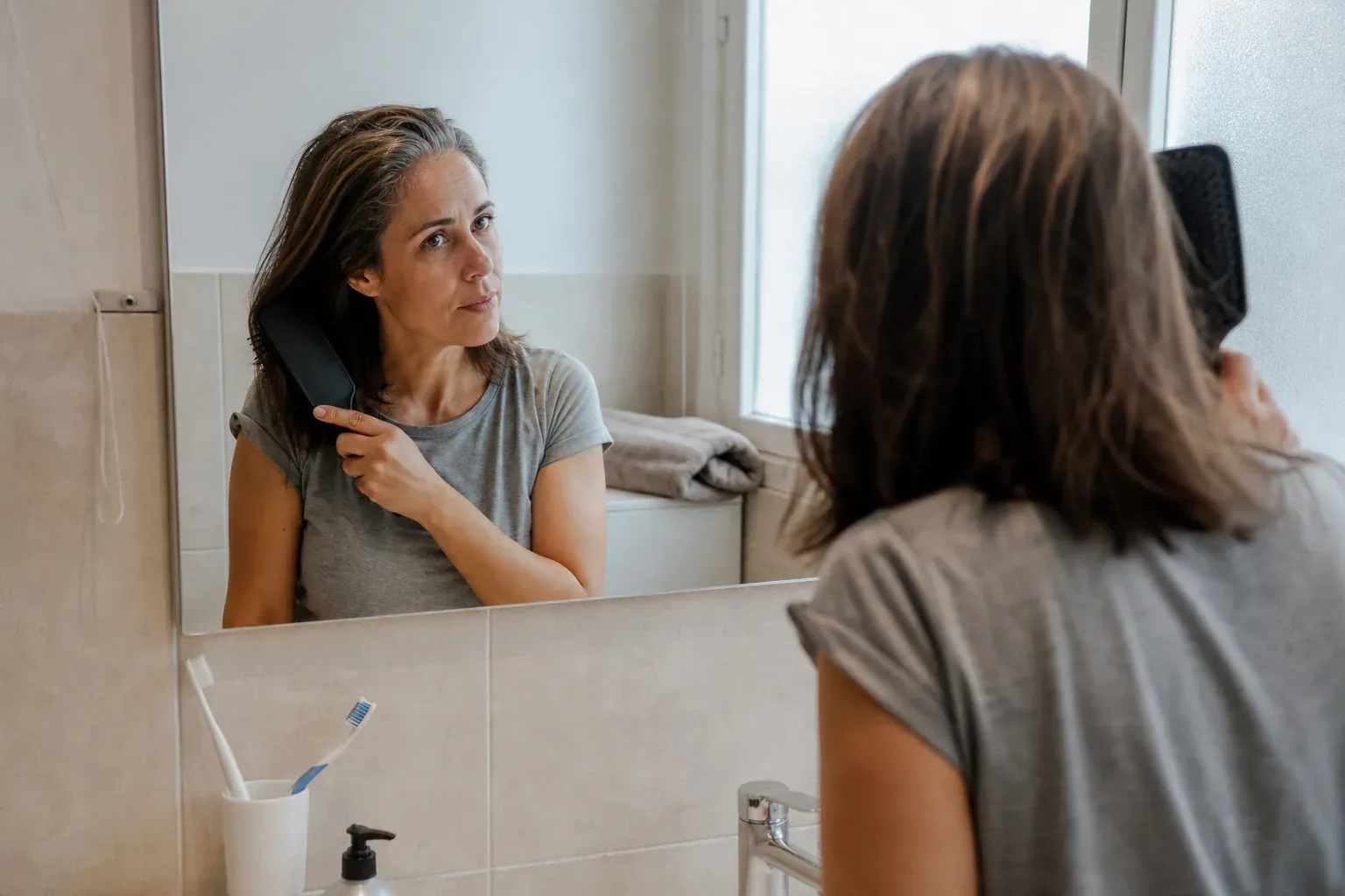 Une femme en tee shirt se coiffe devant le miroir de sa salle de bains, dans la lumière du matin