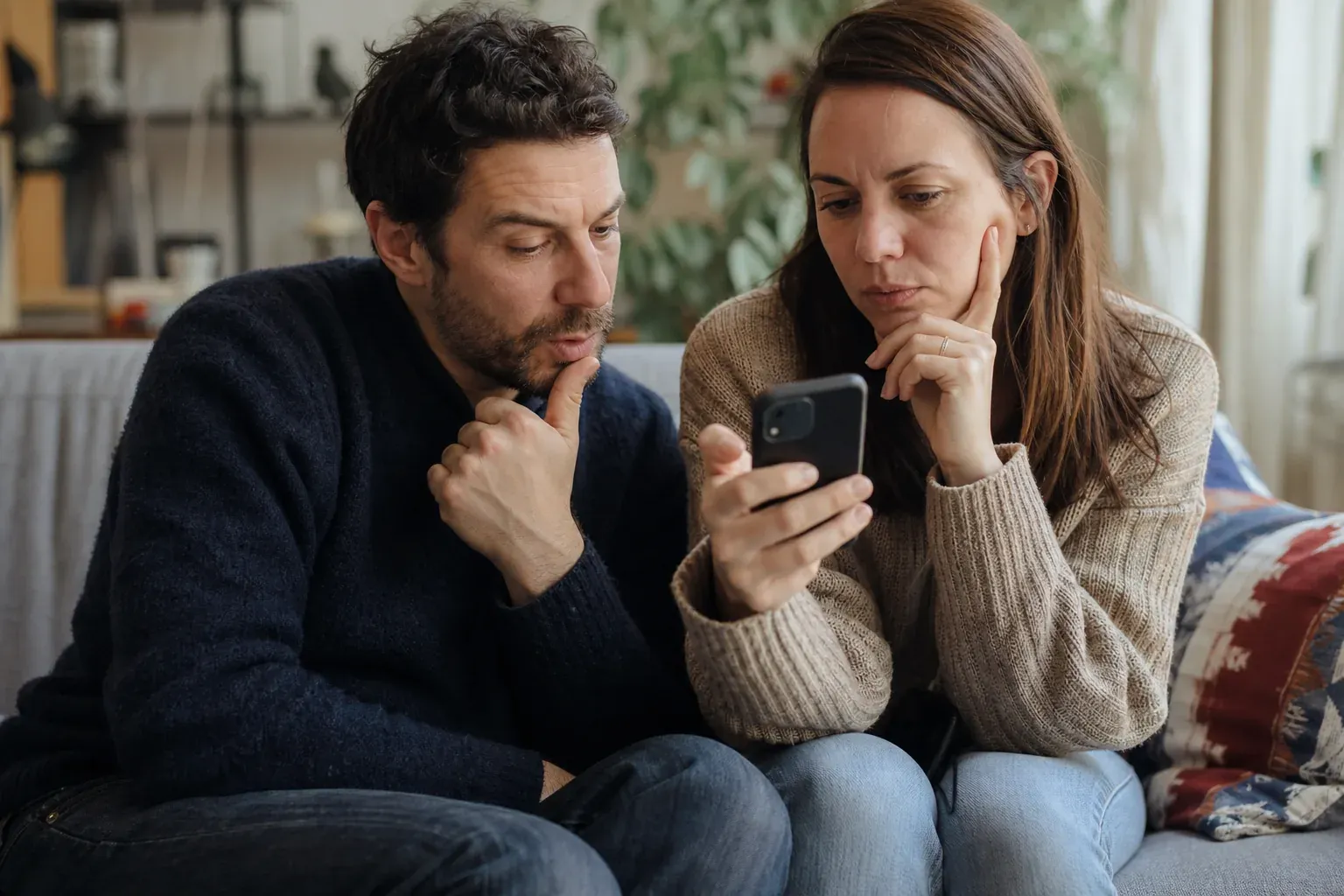 Deux personnes regardent ensemble l'écran d'un téléphone sur un canapé, expression concentrée, salon lumineux